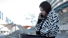 Concentrated brunette lady talking on phone and looking at laptop. Confident business consultant sitting with laptop and talking with interlocutor through phone. Communication and technology concept