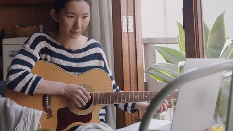 young asian woman playing guitar at home aspiring musician learning to play musical instrument using laptop enjoying creative expression practicing music