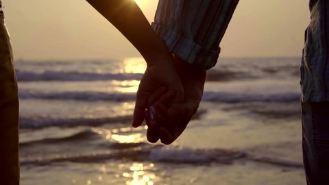 Close-up shot of a couple holding hands and enjoying beautiful sunset on beach, incredible horizon.
