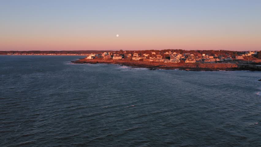 York, Maine / United States - 12 23 2018: Aerial SLIDE to the right past Nubble Lighthouse in Maine lit by a cold December sunrise with a full moon in the distance