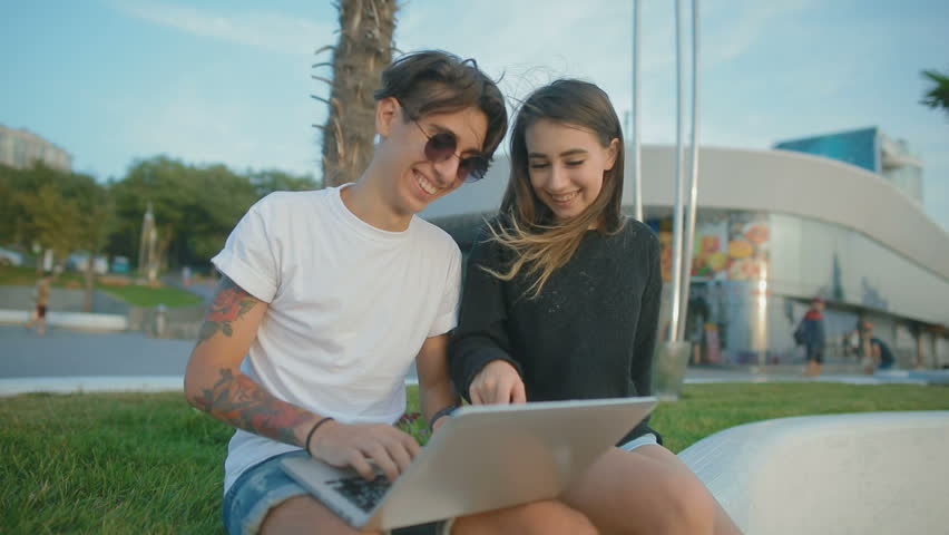 slow motion young smiling man and woman sit on green flowerbed concrete parapet and type on laptop near public building