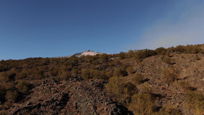 Aerial ascending view over lava rock and shrubs revealing the summit of mount Etna in Sicily (Italy)