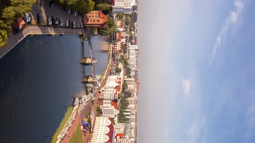 Long transition from day to night. Kaliningrad city center viewed from above, with the Pregolya River flowing through the frame and the Jubilee pedestrian drawbridge connecting the riverbanks. The scene captures the Fishing Village, a modern ethnographic quarter built in German half-timbered style with steep roofs and narrow facades, standing in contrast to surrounding Soviet-era and contemporary structures. The time lapse compresses a full day into seconds: boats move across the water under bright midday sun, shadows lengthen as afternoon fades, the sky shifts through sunset hues, and the district gradually illuminates with warm streetlights and building glow against deep blue twilight. Reflections shimmer on the river surface throughout the transition, and clouds streak across the sky in accelerated motion. The elevated perspective emphasizes the geometric layout of the embankment, the bridge structure, and the dense urban fabric along the waterfront. The Jubilee Bridge opened in 2005 on the preserved pillars of the historic Imperial Bridge destroyed during World War II, marking the city