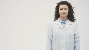 Beautiful young brunette doctor wearing medical clothes on gray background. During this she wears the phonendoscope on her neck. Has beautiful black long hair. Dressed in glasses Slow video.