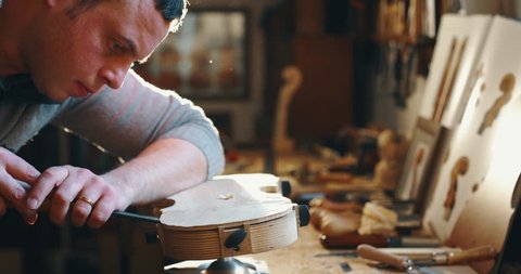 Slow motion close up of master artisan luthier painstaking detailed work on wood violin in a workshop. Shot in 8K. Concept of spiritual instrument, handmade, art, orchestra, artisan, passion for music