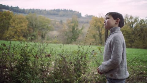 Young playful boy throwing a bright orange into the air as he's walking around a green yard, with view of rural Tuscany in background. Wide shot on 8k helium RED camera.