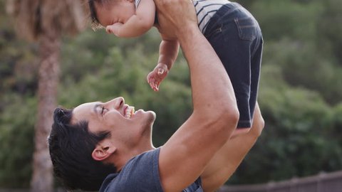 Millennial Hispanic man lifting his baby son in the air in the park, close up