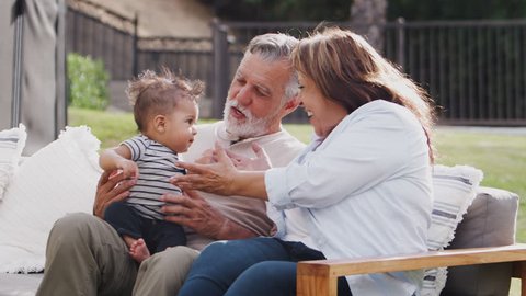 Senior Hispanic couple sitting on a seat in the garden passing their baby grandson over, close up