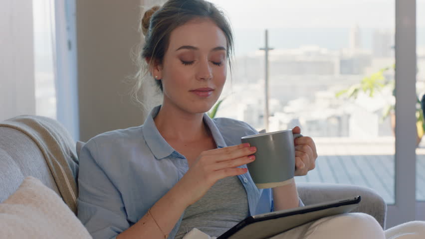 beautiful young woman using digital tablet computer relaxing on couch at home browsing online drinking coffee enjoying comfortable lifestyle