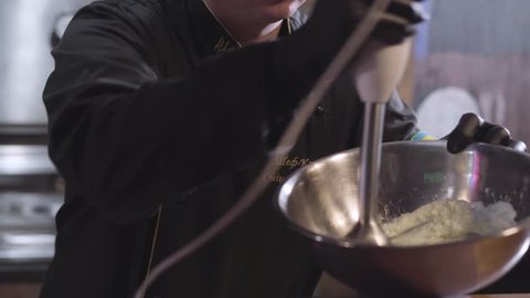 Chef in uniform and black rubber cook gloves whipping sause with blender in big aluminium bowl close up. Preparing food in modern restaurant