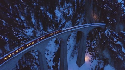 Landwasser Viaduct with Railway and Train at Winter Evening. Swiss Alps, Switzerland. Mountains, Forest and River. Aerial View. Drone Flies Forward, Camera Tilts Down