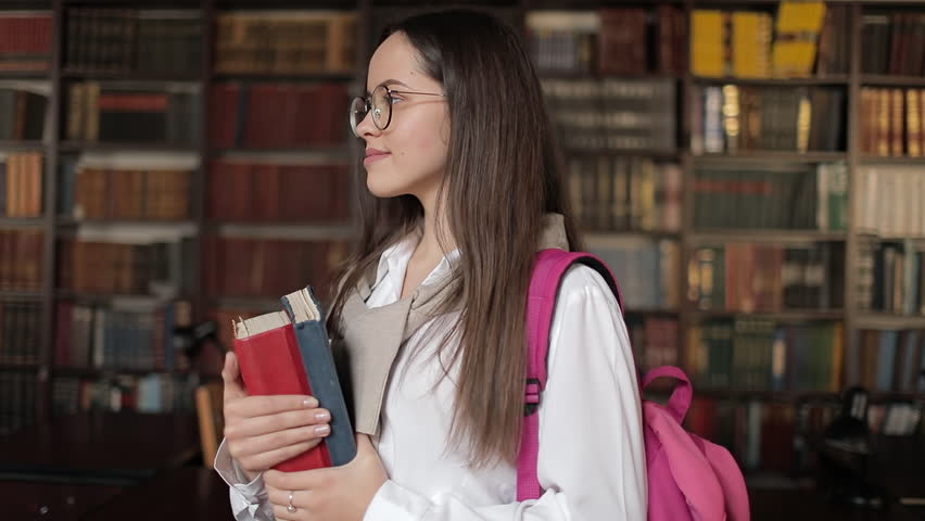 Girl holding books in her hand in library and smiling toward camera