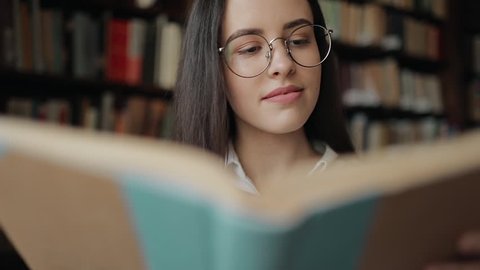 Smiling young girl in glasses reading book in library of university