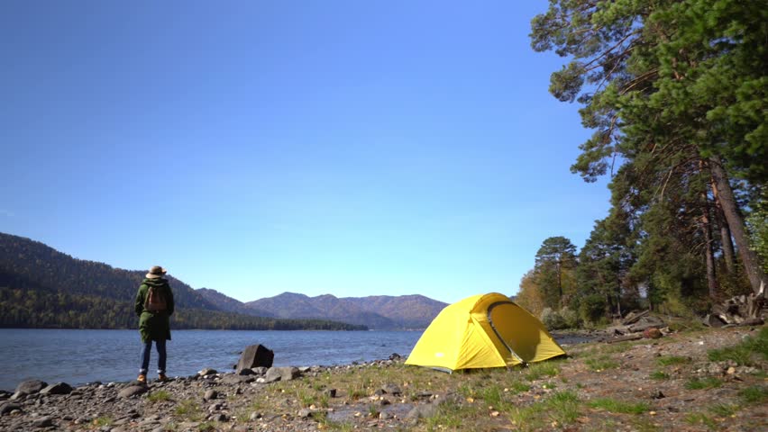 The woman the traveler goes on the coast of the mountain lake. On the bank of the lake there is a yellow tent.