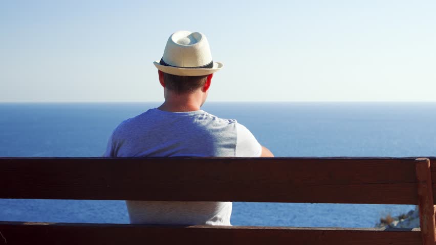 Young handsome man sitting relaxing on bench at edge of cliff take pictures on cellphone. Male in hat enjoying breathtaking view of blue Mediterranean sea making photos on mobile phone