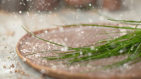 SLOW MOTION, DOF, MACRO: Water drops splash over the wooden table after homegrown chives fall into a plate full of refreshing cold water. Organic long green chive stalks crash into a brown plate.
