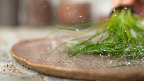 SLOW MOTION, MACRO, DOF: Unrecognizable person splashing water by shaking a pot of chives in a shallow stone plate. Blurry human holding a pot of organic chives and spraying water over the table.
