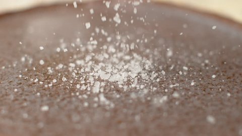 SLOW MOTION, DOF, CLOSE UP: Coarse crystals of rock salt are scattered across the granite countertop. Cinematic shot of white crystals of sugar bouncing off the hard gray stone surface of the table.