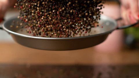 MACRO, DOF, SLOW MOTION: Peppercorns flying around the kitchen as young woman roasts them in a hot metal pan. Unrecognizable female chef toasting aromatic and colorful peppercorns in a frying pan.