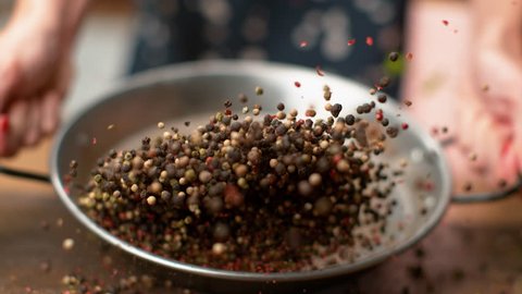 SLOW MOTION, MACRO, DOF: Unrecognizable woman toasting colorful peppercorns in a metal pan. Female chef mixing fragrant multicolored pepper seeds in a frying pan. Peppercorns flying around kitchen.