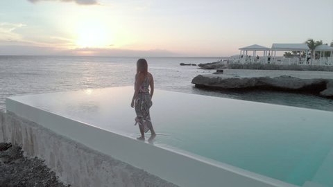 Aerial Backward: Woman Walking on the Edge of Swimming Pool next to the Ocean