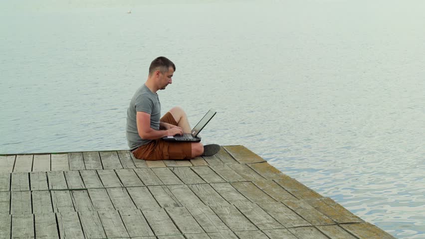 Handsome man in casual wear sitting on a wooden bridge over the blue water and works on a laptop. Male on an old pier working by the beautiful river