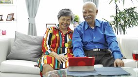 4K Asian senior couple having fun video chat on tablet mobile. Sharing social media communication sitting on sofa at home. Slow motion dolly shot.