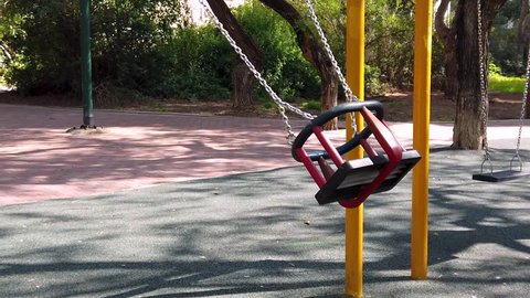 Slow motion shot of an empty swing, swinging alone in a playground without children. The trees in the background cast shadows over the new safety matt and the old brick road. Steady handled.