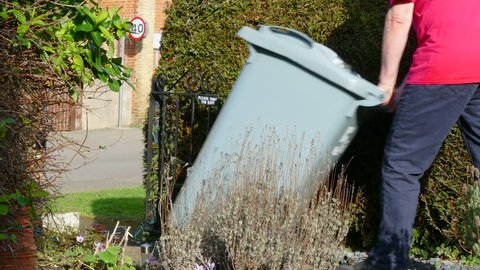 Man quickly pushing a large plastic wheelie bin full of domestic waste, down the path of his garden, out through the gate and onto the street, to leave the trash / refuse ready for collection.