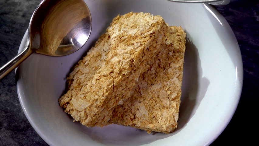 Slow motion close POV overhead shot of steaming hot milk being poured from a small jug onto a ceramic bowl containing dry wheat biscuits cereal.