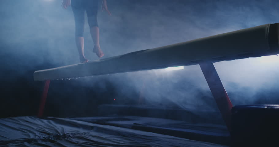 The girl is a professional athlete performs gymnastic acrobatic trick on a beam in backlight and slow motion in sports gymnastic clothing. Smoke and blue. Jump and spin on the balance beam
