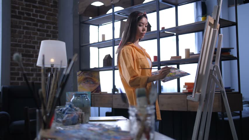 Inspired lovely female artist with colored palette creating artwork in workshop. Professional woman painter with paintbrush and palette drawing picture on canvas, expressing visions and imagination. | Shutterstock HD Video #1024666712