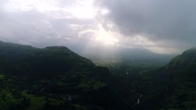 Aerial shot of early morning sun rays reaching the green surface of the mother earth passing through the clouds, surrounded by beautiful green mountains.