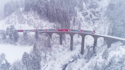 GRAUBUNDEN, SWITZERLAND - FEBRUARY 11, 2019: Viaduct and Glacier Express Train in Winter Day. Snowing. Swiss Alps. Switzerland. Aerial View. Drone Follows Train