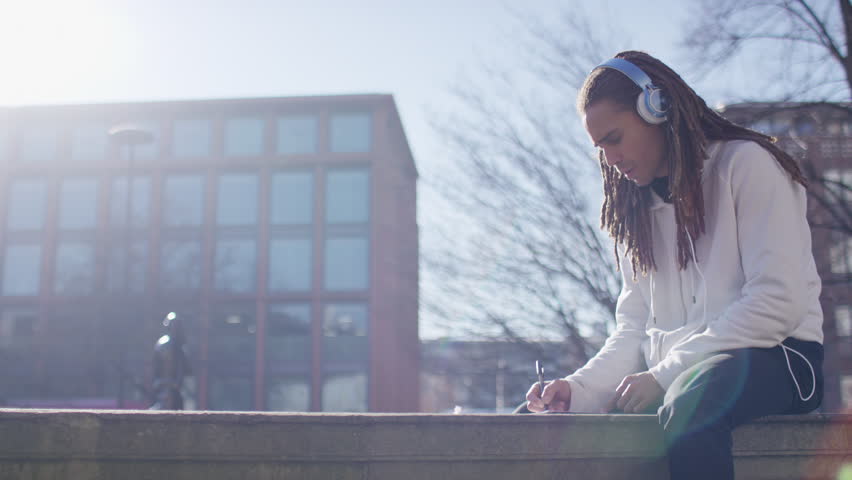Handsome man with dreadlocks writing and listening to music as he sits on a wall