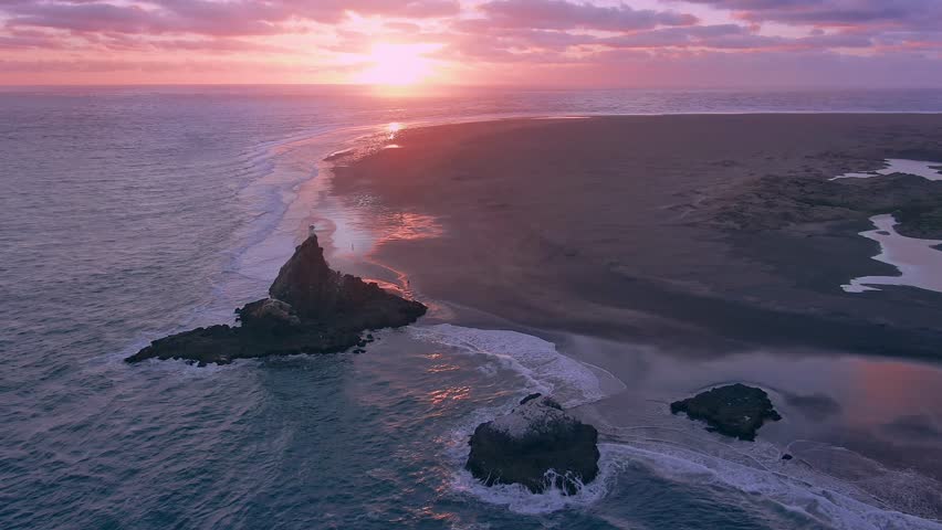 Aerial: Sun setting over beach and pink sky of the rocky coastline of Whatipu sunset. The beach is situated in the Waitakere Ranges at the opening of the Manukau Harbour. Auckland, New Zealand. 