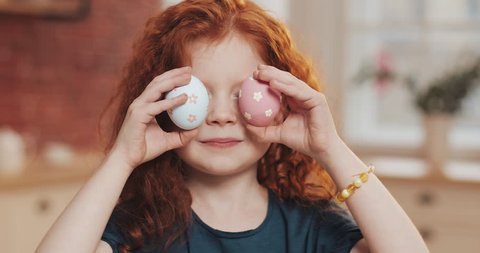 Portrait of cheerful redhead little kid girl playing with easter egg on the kitchen background. She is cheering and having fun at the camera. Happy Easter
