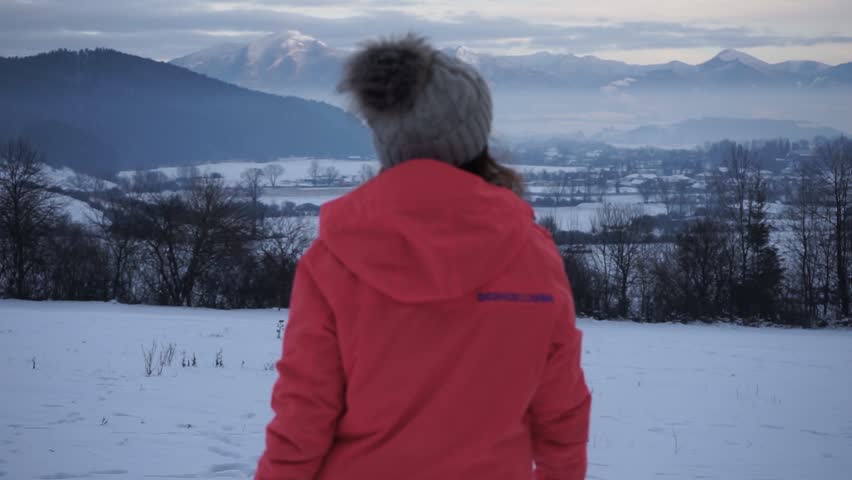 Young girl in an orange winter jacket happily walking in a winter snowy meadow with a view of mountains