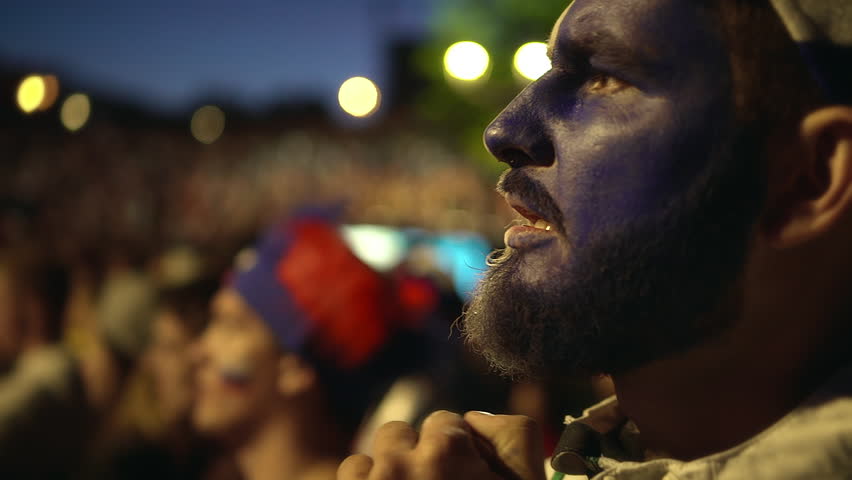 French fan with paint on his face intensely watching football game, experiencing. Man hold his hand in face of excitement sport match. guy worried against background crowd from loss his favorite team
