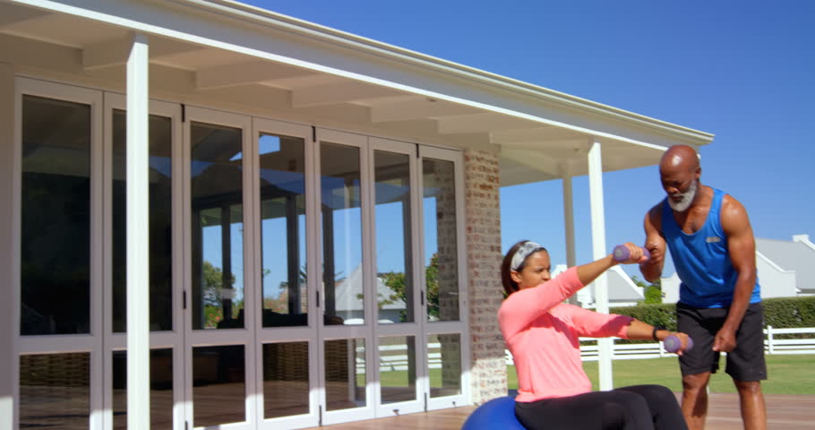 Mature man assisting mature woman in exercise at backyard on a sunny day. She is using dumbbells in the sunshine. Social distancing and self isolation in quarantine lockdown for Coronavirus Covid19