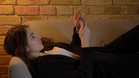 Portrait of young caucasian girl with wavy hair lying on sofa and watching into smartphone in cosy home atmosphere.