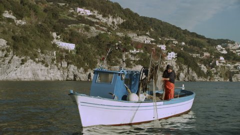 Two busy fishermen sailing in a boat along the ocean with a view of the Amalfi Coast in the background on a bright sunny day. Wide shot on 8k helium RED camera.