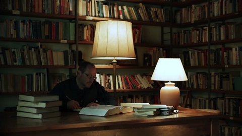 Hardworking Italian man reading and writing at a desk with piles of books on top with bookshelves of books in the background. Medium shot on 8k helium RED camera.