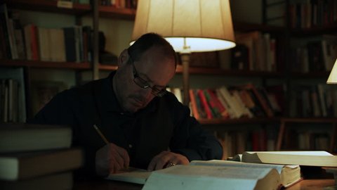 Hardworking Italian man reading and writing at a desk with piles of books on top with bookshelves of books in the background. Medium shot on 8k helium RED camera.