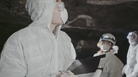 Person in bio-hazard suit, face mask and goggles wearing a headlamp standing taking notes in an underground tunnel as members of the team move around in the background.