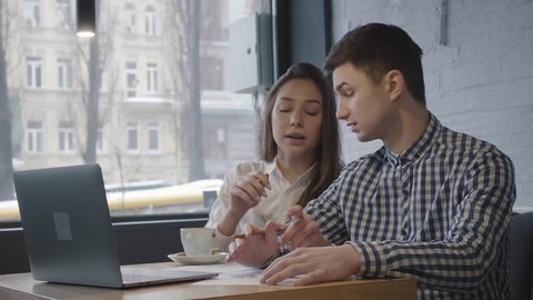 Young couple working in modern cafe together close up. Confident man explaining to girl information and writes it on paper. Woman listening to partner and corrects what was written. Laptop standing on