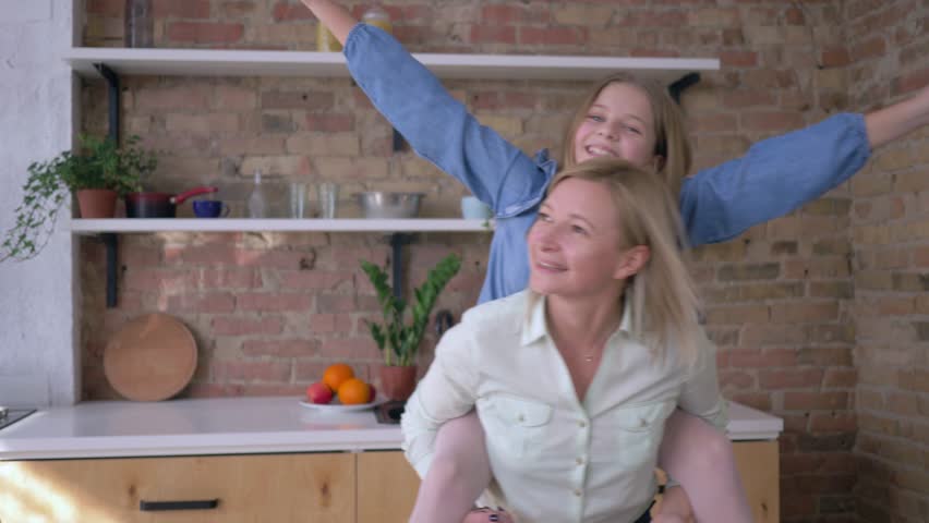 games with mother, funny little girl plays with mom sitting on back and makes plane of arms extended in family kitchen at home