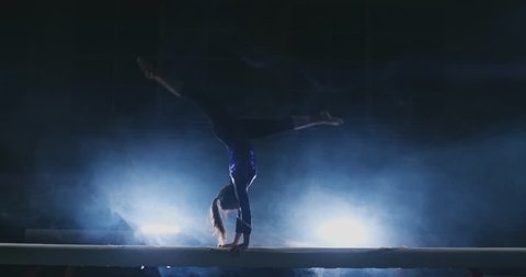 The girl is a professional athlete performs gymnastic acrobatic trick on a beam in backlight and slow motion in sports gymnastic clothing. Smoke and blue. Jump and spin on the balance beam