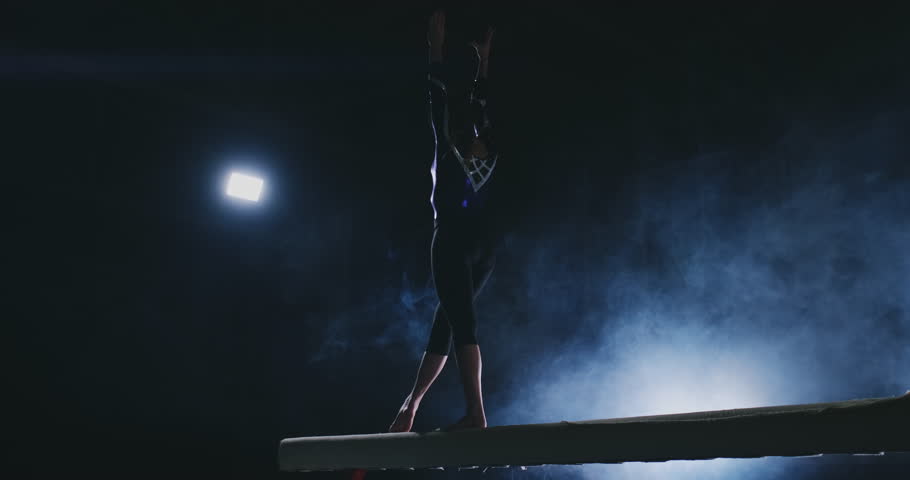 The girl is a professional athlete performs gymnastic acrobatic trick on a beam in backlight and slow motion in sports gymnastic clothing. Smoke and blue. Jump and spin on the balance beam