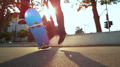 SUN FLARE, CLOSE UP, SLOW MOTION: Unrecognizable skateboarder lands a difficult fakie flip while cruising through the modern city on sunny day. Extreme young man skateboarding and doing cool tricks.
